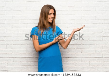 Woman with blue dress over brick wall extending hands to the side for inviting to come