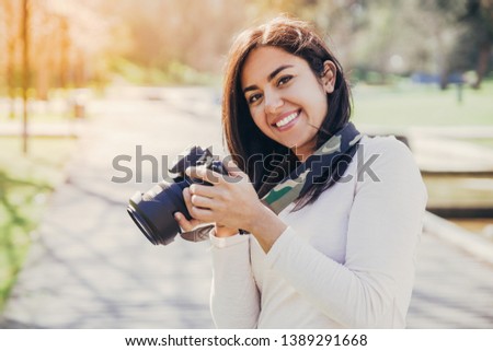 Positive successful photographer enjoying photo-shooting outdoors. Young woman in casual holding photo camera and smiling for portrait. Hobby concept