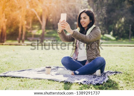 Excited student girl resting in park and taking selfies for social media. Young woman in casual sitting on grass, using smartphone and enjoying coffee break. Communication concept