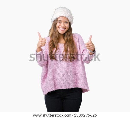 Young beautiful brunette woman wearing sweater and winter hat over isolated background success sign doing positive gesture with hand, thumbs up smiling and happy. Looking at the camera 