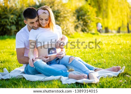 Happy couple expecting baby, pregnant young woman with husband sitting on green grass and holding ultrasound pictures of baby, young family and new life concept