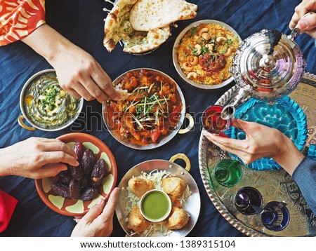 Ramadan iftar meal theme people eating variety foods together on dining table with large dates (Medjool), Samosas, Aloo Gobi, Chicken Biryani, Moroccan tea set, Palak Paneer and Naan bread. (top view) Royalty-Free Stock Photo #1389315104