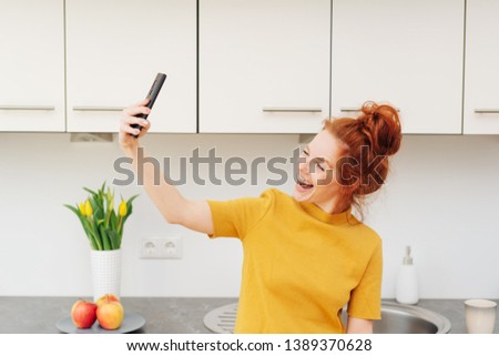 Laughing vivacious young redhead woman taking a selfie on her mobile phone indoors in her kitchen