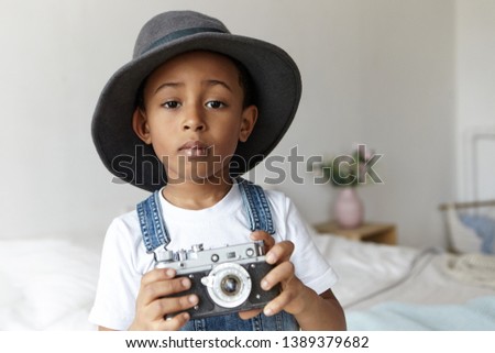 People, photography, art and hobby concept. Indoor image of cute adorable Afro American boy posing in cozy bedroom interior wearing stylish round hat, holding vintage camera, taking pictures