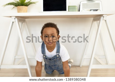 Picture of handsome cute Afro American boy of preschool age posing in stylish apartment interior, looking at camera with curious or suspicious look, being home alone. Selective focus on face