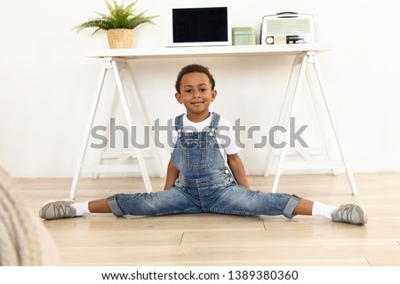 Horizontal shot of active energetic little African American boy wearing denim jumpsuit and running shoes exercising at home, doing side splits on floor. Fitness, sports, activity and childhood