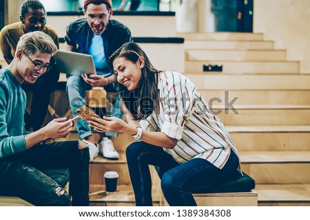 Cheerful millenial woman showing latest feed from social networks to group of multiracial friends, happy crew of male and female colleagues spending free time in college talking and using gadgets Royalty-Free Stock Photo #1389384308