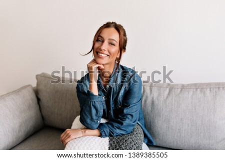 Happy laughing girl with tanned skin and white teeth sitting at sofa at cosy apartment and posing at camera 