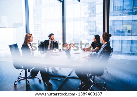 Concept of brainstorming and briefing, positive diverse male and female financial experts discussing information during working time in conference room sitting at desktop with laptop devices Royalty-Free Stock Photo #1389400187