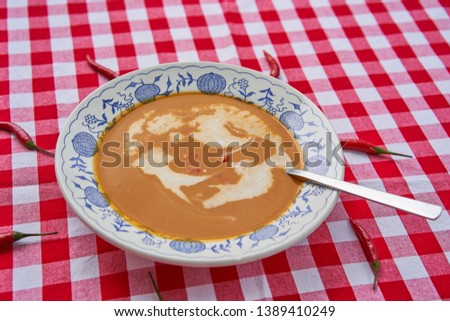 Spicy hot asian style sweet potato or yam creamy soup with coconut milk served in rustic deep plate with spoon on the table covered by checkered red and white tablecolth with small red chili peppers.