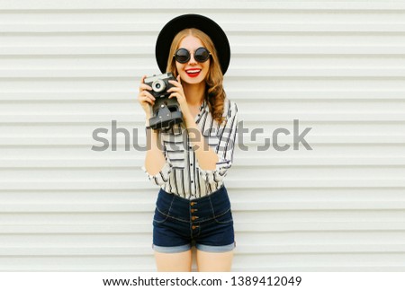 Happy smiling young woman holding vintage film camera in black round hat, shorts, white striped shirt on white wall background