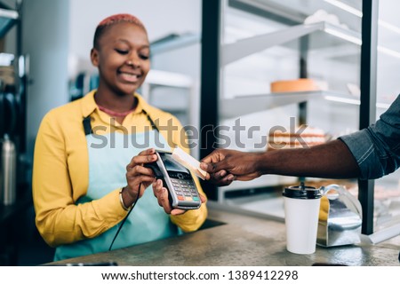 Smiling African American woman in apron smiling and holding payment terminal for anonymous customer paying with credit card for food in cozy cafe