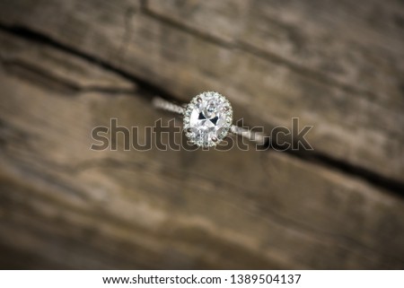 Elegant and simple womens engagement diamond ring with a oval halo setting on a gray brown wood barnwood blurred background nestled in the crack between two planks 