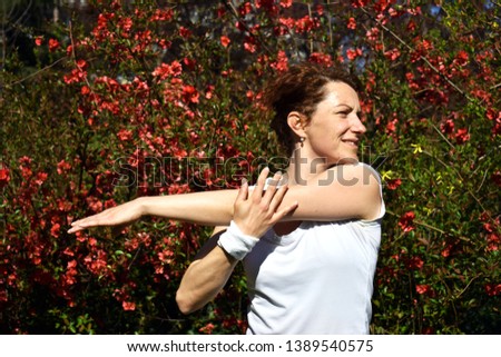 Red hair girl is stretching her arm after trainin in city park, bush with red flowers in background