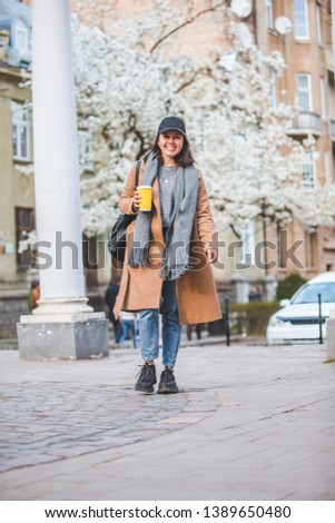 young stylish woman walking by street with coffee cup. spring blooming tree on background