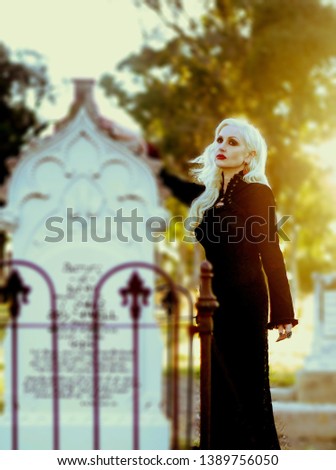 Photo of a pretty blonde woman dressed in a long black dress standing next to an old gravestone 