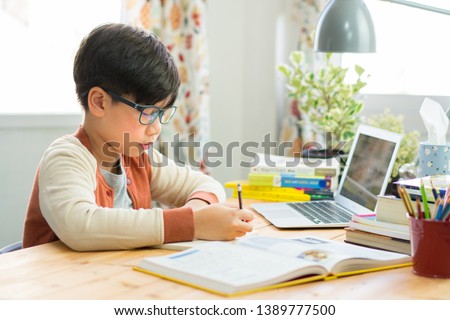 A smart preteen boy sitting at the table, writing homework or preparing for the exam. Teen using notebook computer to study. Student, School, Remote, Online learning, New normal, Home Based Learning. Royalty-Free Stock Photo #1389777500