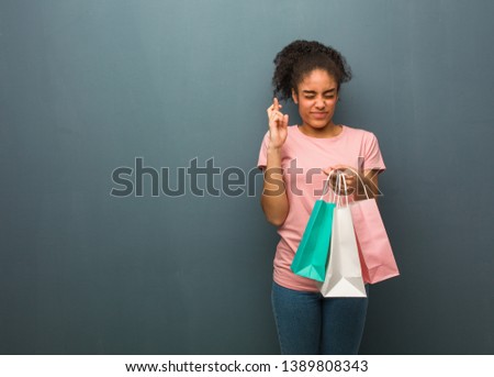 Young black woman crossing fingers for having luck. She is holding a shopping bags.
