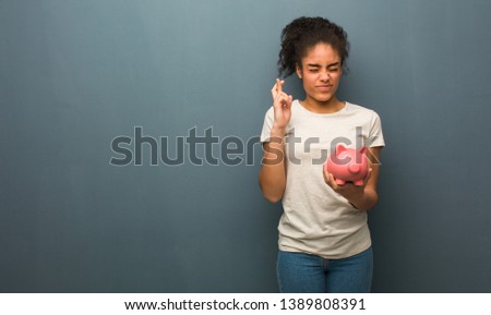 Young black woman crossing fingers for having luck. She is holding a piggy bank.