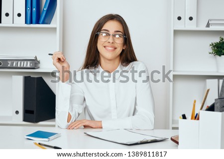Happy woman in shirt and glasses is sitting at the table in the office and smiling in the camera folder with documents pencils pens                    