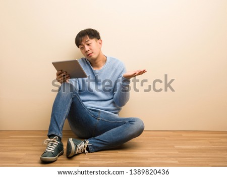 Young chinese man sitting using his tablet doubting and shrugging shoulders