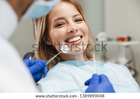 Image of satisfied young woman sitting in dental chair at medical center while professional doctor fixing her teeth Royalty-Free Stock Photo #1389890048