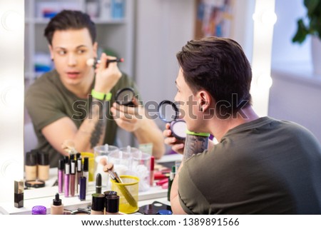 Peaceful young man. Curious handsome guy getting ready in front of mirror and applying layer of powder