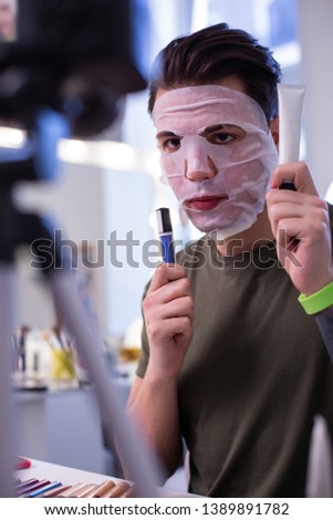 Handsome professional blogger. Resolute neat guy wearing wet tissue mask during video recording and carrying tubes with products