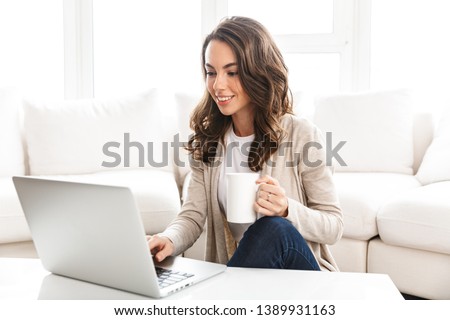 Beautiful young woman working on laptop computer while sitting at the living room, drinking coffee Royalty-Free Stock Photo #1389931163