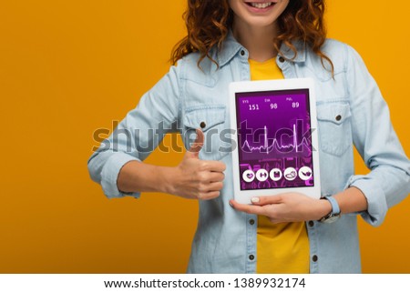 cropped view of cheerful curly girl holding digital tablet with e health app screen and showing thumb up isolated on orange 