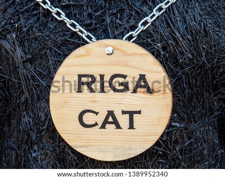Wooden round sign on a metal chain, Riga cat is written onthe sign.Black wooden branches background.