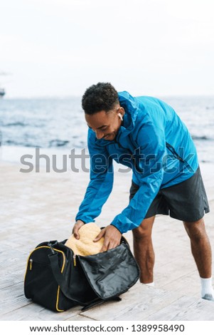 Picture of a handsome young african sports man outdoors at the beach sea walking with bag listening music with earphones.