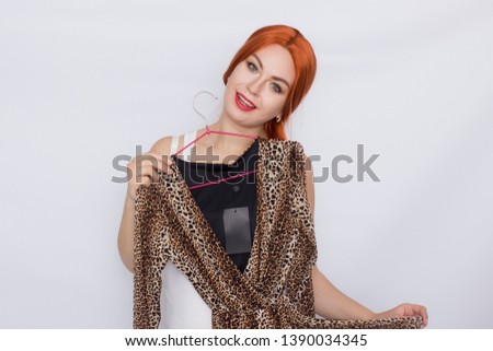 Photo of beautiful redhead woman in white shirt holding dress with leo print over white background in studio
