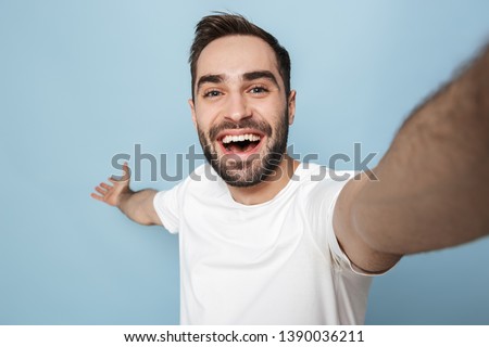 Cheerful excited man wearing blank t-shirt standing isolated over blue background, taking selfie