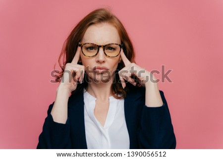 Portrait of intense beautiful lady touches temples with both hands, tries to recollect something important in mind, has serious concentrated look at camera, wears spectacles and elegant suit