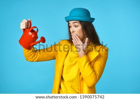 Beautiful woman in a yellow jacket in a blue hat holding a watering can in her hands a blue background.