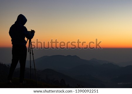 silhouette of Travel photographer standing with a camera mounted on a tripod and shooting a time lapse of the sunrise/sunset. man wearing his hood enjoying the mountain view /valley view.