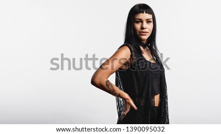 Woman with fashion piercing on lips standing against white background with hand on waist. Portrait of a young gothic woman with tattoo on hand looking at camera.