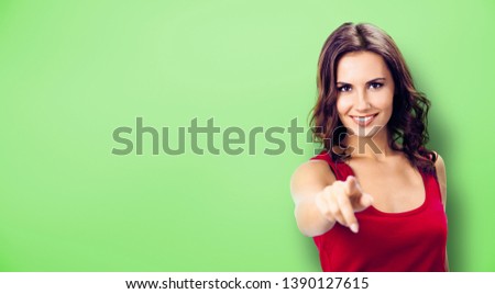 Pointing at You - advertising concept photo. Smiling woman in red, showing copy space, visual imaginary or something, or pressing virtual button, green background. Copyspace for some text or slogan. 