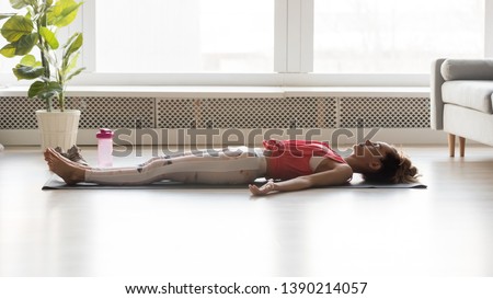 Side view full length image of woman wearing activewear lying on yoga mat practicing corpse pose resting after working out at home, savasana exercise, body care, yogi female healthy lifestyle concept