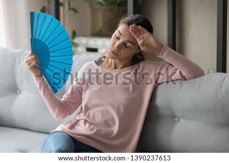 Overheated female sitting on couch in living room at hot summer weather day feeling discomfort suffers from heat waving blue fan to cool herself, girl sweating dwelling without air conditioner concept Royalty-Free Stock Photo #1390237613