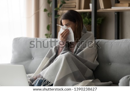 Sick young woman sitting on couch at home, unhealthy girl covered with warm plaid sneezing blowing her nose on paper tissue suffering from grippe temperature or seasonal allergy, cold dwelling concept Royalty-Free Stock Photo #1390238627