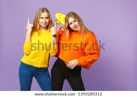 Two beautiful young blonde twins sisters girls in vivid clothes pointing index finger up hold yellow skateboard isolated on violet blue background. People family lifestyle concept. Mock up copy space