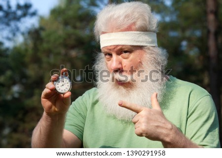 Joyful senior sportive man pointing on stopwatch. Outdoor lifestyle portrait against natural tree background. Active leisure.