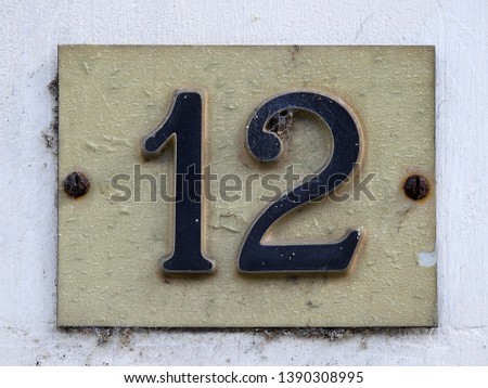 House number twelve (12) stylish sign black on white plate against white wall background (closeup). Grunge, texture, wallpaper