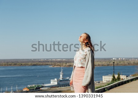 Beautiful woman with long hair enjoying the city view from the bridge on a Sunny day. Beautiful brunette in a knitted jacket, spending time on the waterfront while relaxing in Europe.
