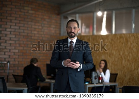 Young Beardy Businessman Holding Phone While Standing Middle Office. Having Colleagues On Background,