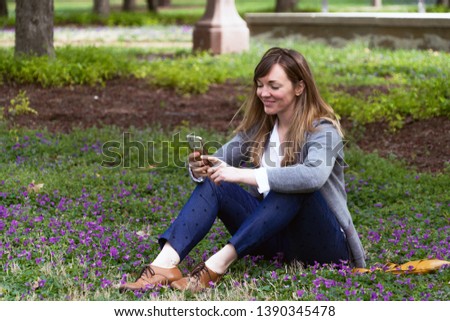 A young, classy, attractive woman sitting on the grass in the park smiling as she looks at her phone texting or reading  - looking very natural seems happy- like an ad for a lifestyle brand