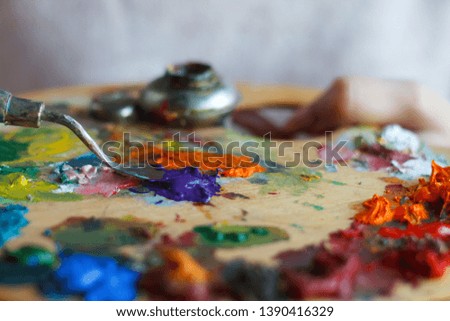 Close-up of female hands mixing paints on a palette with a spatula against a white canvas, creating an oil painting, soft focus