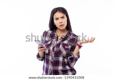 Portrait of female student holding mobile phone in right hand and looking at the camera with shocked expression on her face, wearing casual plaid shirt, isolated on white background